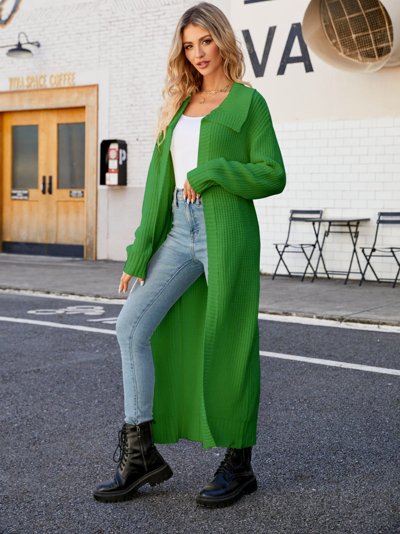 A woman standing on the sidewalk wearing a green collared open front duster cardigan over a white top and blue jeans, with black boots.
