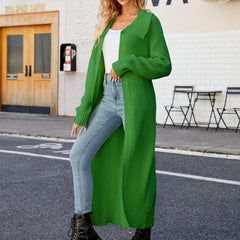 A woman standing on the sidewalk wearing a green collared open front duster cardigan over a white top and blue jeans, with black boots.
