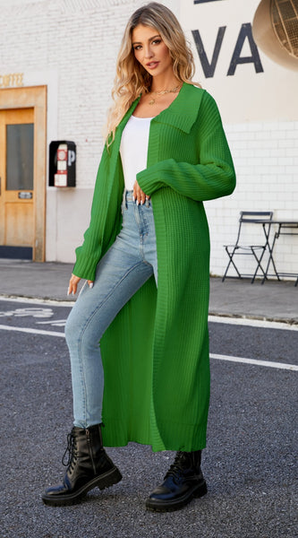 A woman standing on the sidewalk wearing a green collared open front duster cardigan over a white top and blue jeans, with black boots.