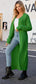A woman standing on the sidewalk wearing a green collared open front duster cardigan over a white top and blue jeans, with black boots.