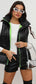 A woman wearing a black zip-up collared vest with green accents and a white top, holding a silver purse, with black boots and green earrings.